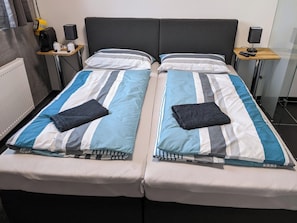 Un llit doble amb roba de llit estriada, dues taules de nit amb làmpades, un radiador i una petita estant amb objectes.