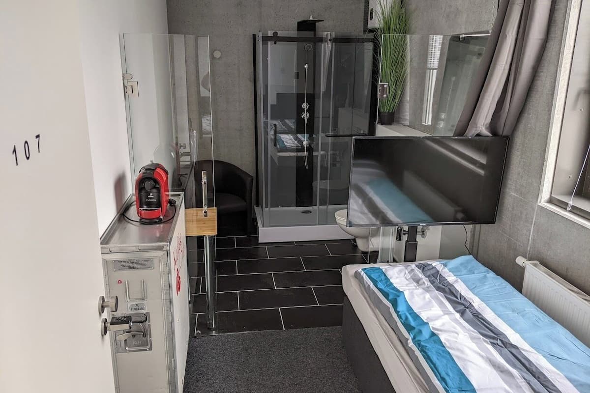 Habitación de hotel compacta con cama, ducha, escritorio pequeño y un dispositivo rojo, y televisor.