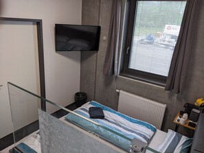 A small hotel room with a bed, a TV mounted on the wall, a window with blinds, a radiator, and a small table with items on it. - Business-motel mit Xl-parking (Heimsheim)