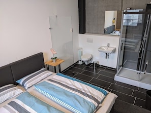 Habitació d'hotel moderna amb llit, taula de nit petita amb una flor, bany amb rentapla i vàter, i dutxa amb envàs de vidre.