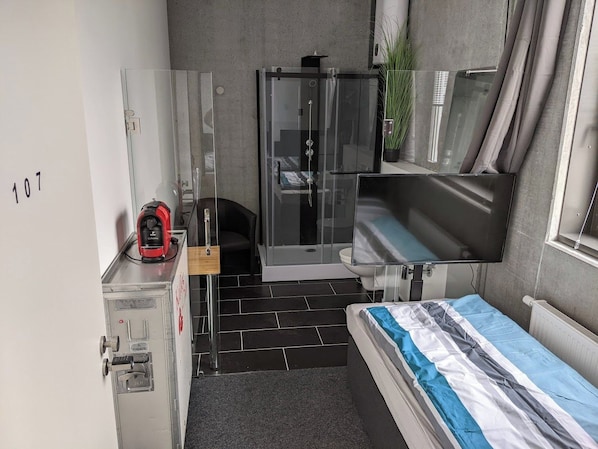 Habitació d'hotel senzilla amb llit, dutxa, tauleta petita amb un dispositiu vermell i televisió.