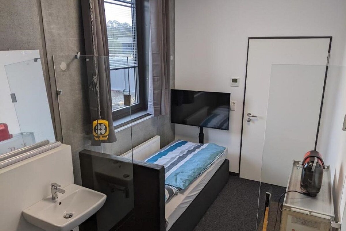 A compact hotel room with a single bed, a bedside TV, a small bathroom with a sink and mirror, and a window with blinds.