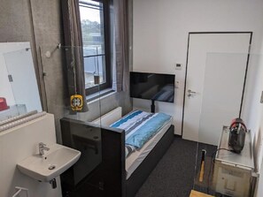 A compact hotel room with a single bed, a bedside TV, a small bathroom with a sink and mirror, and a window with blinds. - Business-motel mit Xl-parking (Heimsheim)