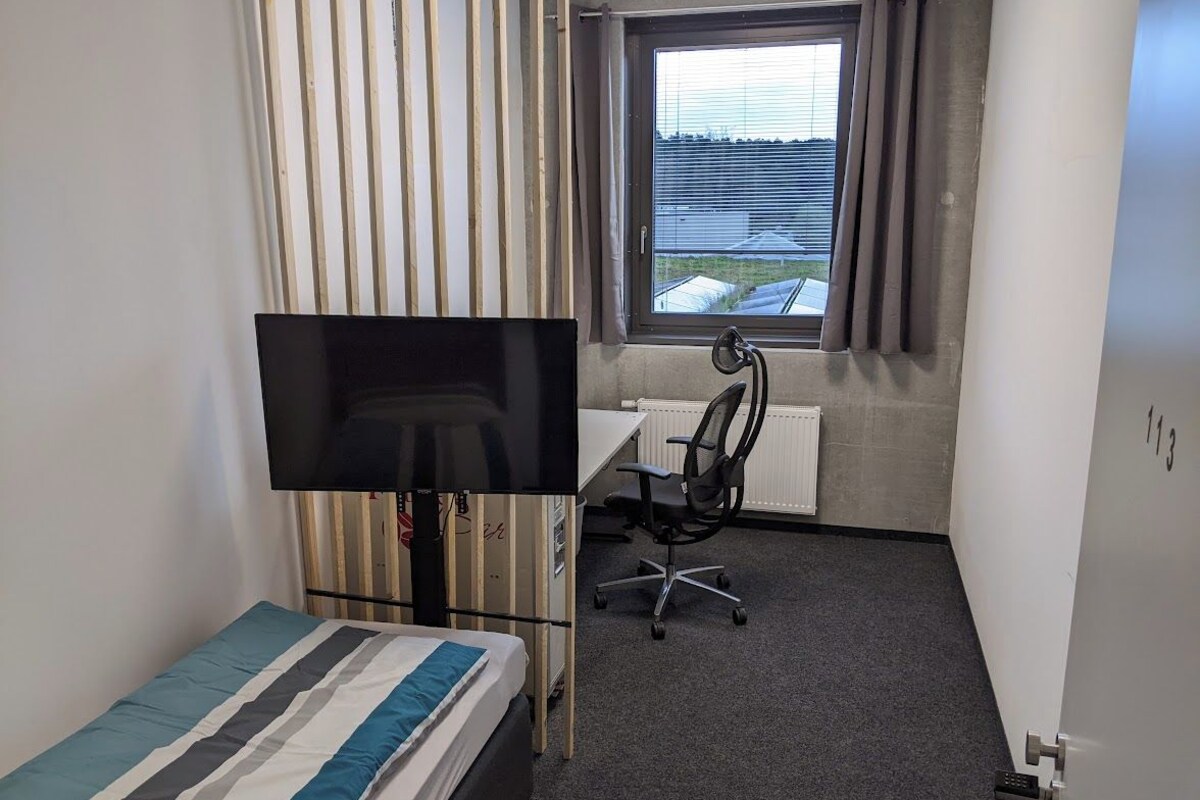 Una habitación pequeña y funcional con cama, escritorio, silla, televisor y ventana con cortinas.