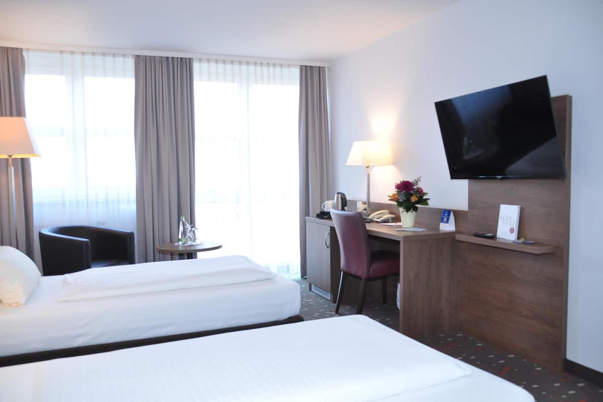 A hotel room with two beds, a desk with a chair, a television mounted on the wall, and a window with curtains.