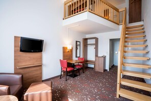 Une chambre d’hôtel avec un escalier en bois, un téléviseur fixé au mur, un coin repas avec une table et des chaises, et une moquette à motifs.