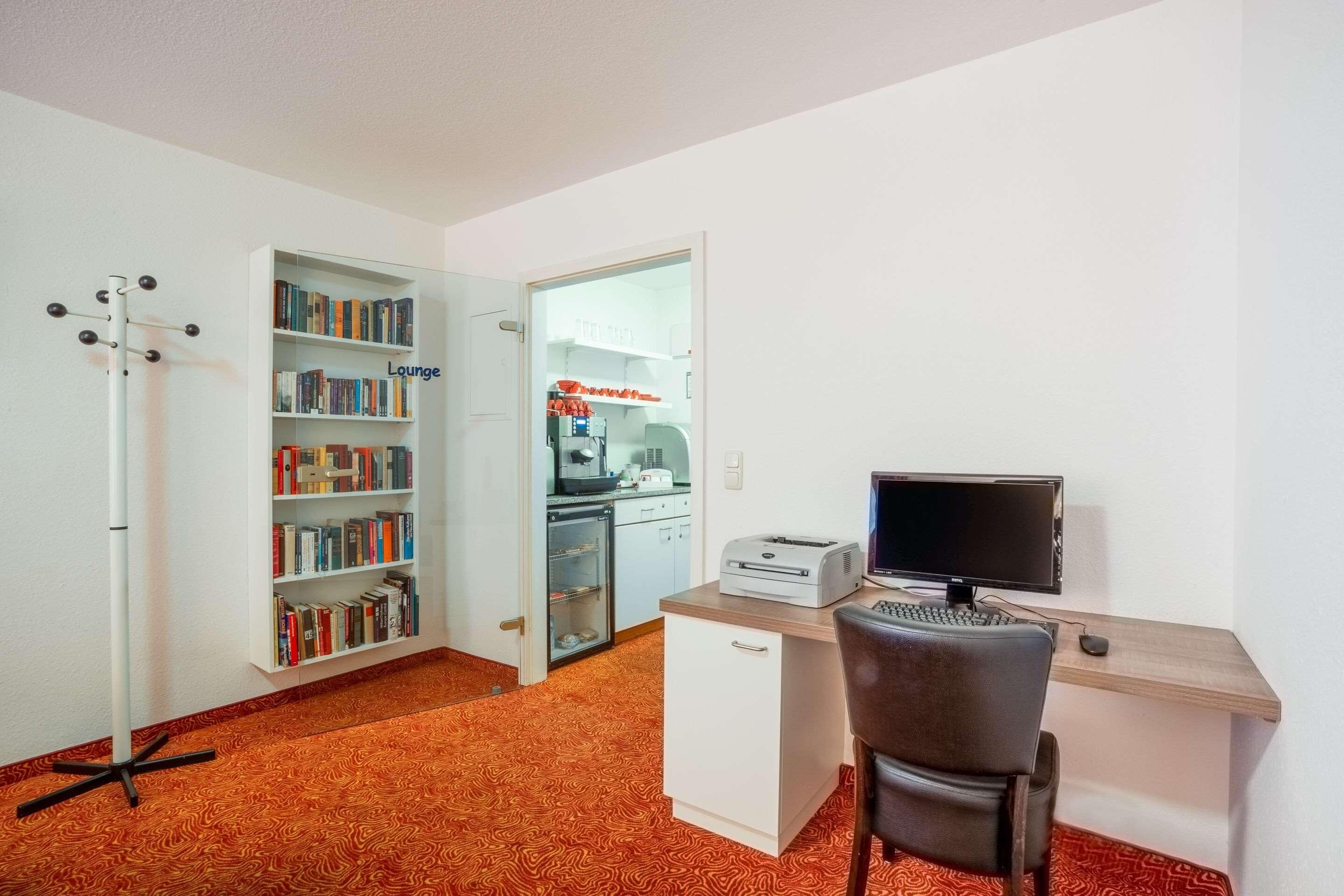 Ein kompakter Arbeitsraum mit Schreibtisch, Stuhl, Computer, Drucker, Bücherregal und einer kleinen Küchenzeile.