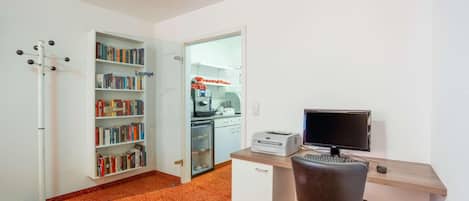 Um espaço de escritório compacto com escrivaninha, cadeira, computador, impressora, estante e área de cozinha compacta.