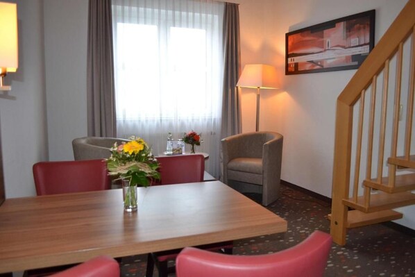Une chambre d’hôtel avec une table à manger en bois, des chaises rouges et grises, un vase avec des fleurs, une lampe et un tableau au mur.