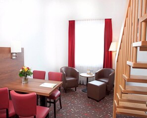 A hotel room with a wooden desk, red chairs, a small round table, and a sofa. There is a staircase to the right and a window with red curtains. - Best Western Hotel Erlangen (Erlangen)