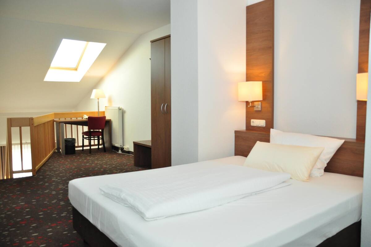 A hotel room with a bed, a desk with a chair, a lamp, and a skylight.