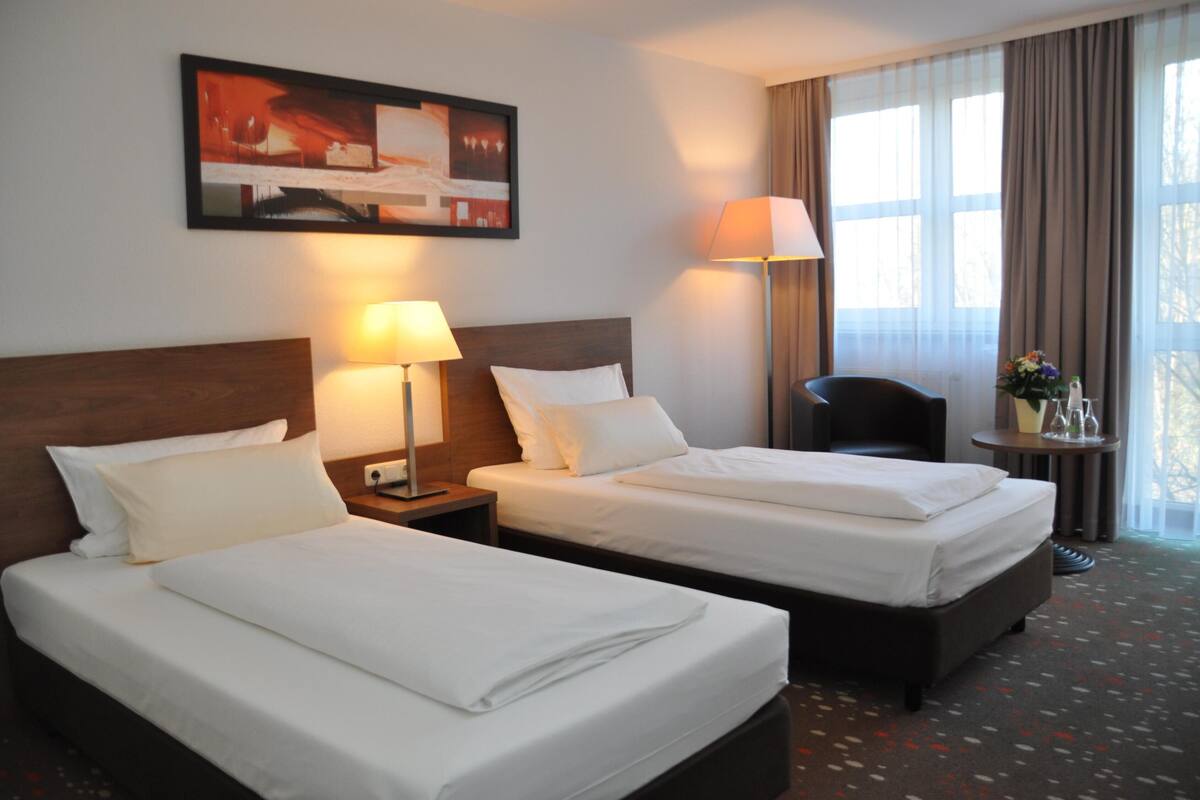 A hotel room with two beds, a desk, a chair, a lamp, and a painting on the wall.