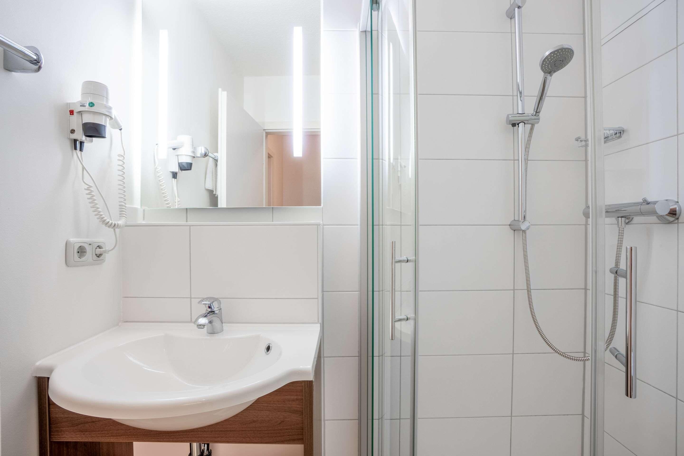 Ein Badezimmer mit einem weißen Waschbecken, einer Duschkabine aus Glas und einem an der Wand befestigten Fön.