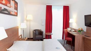Ein Hotelzimmer mit einem Bett, zwei Nachttischen mit Lampen, einem Schreibtisch mit Stuhl, einem Fernseher und einem Fenster mit roten Vorhängen.
