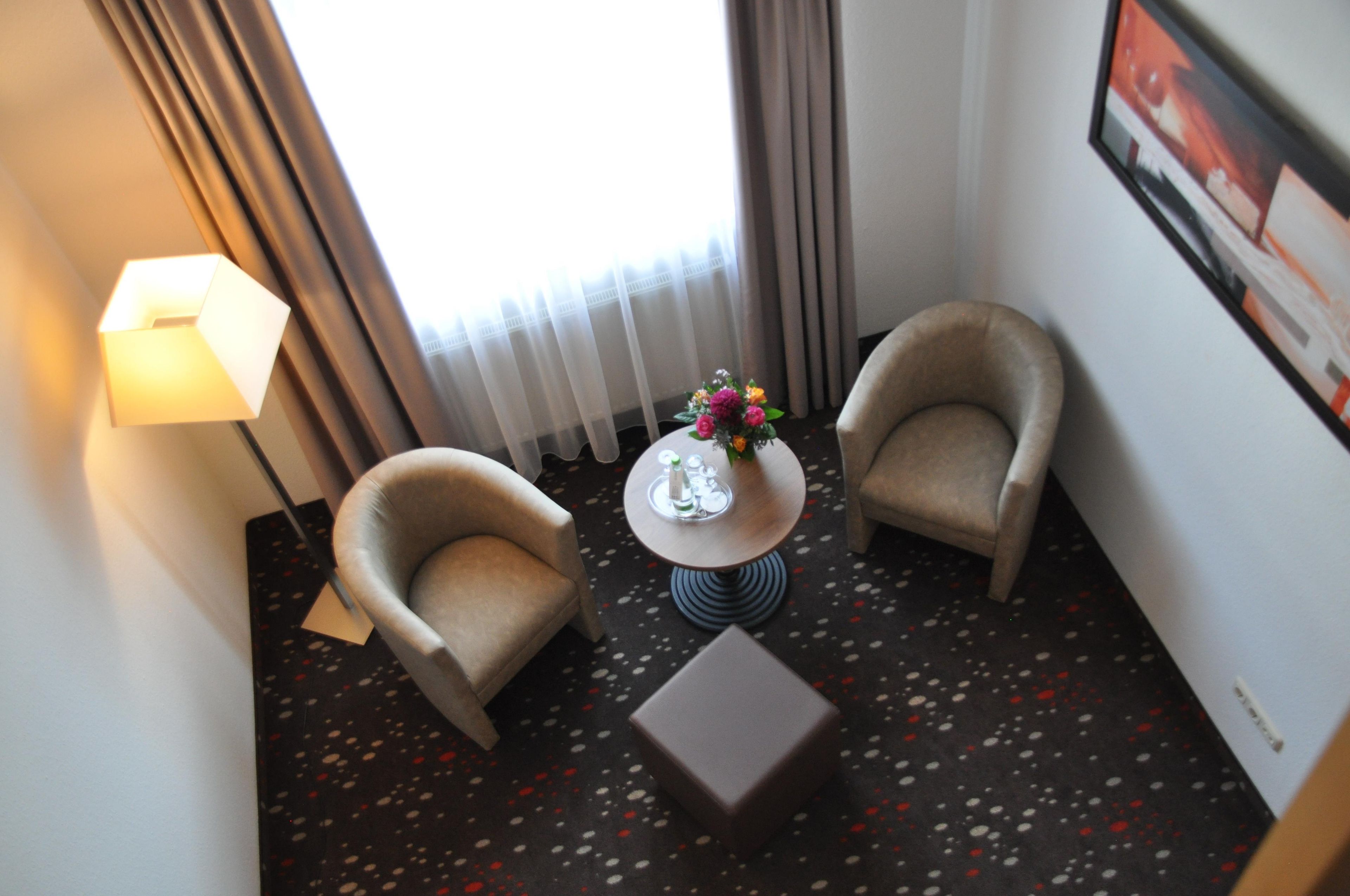 Ein Hotelzimmer mit zwei Sesseln, einem kleinen runden Tisch mit Blumenvase, einer Lampe und einem gerahmten Bild an der Wand.