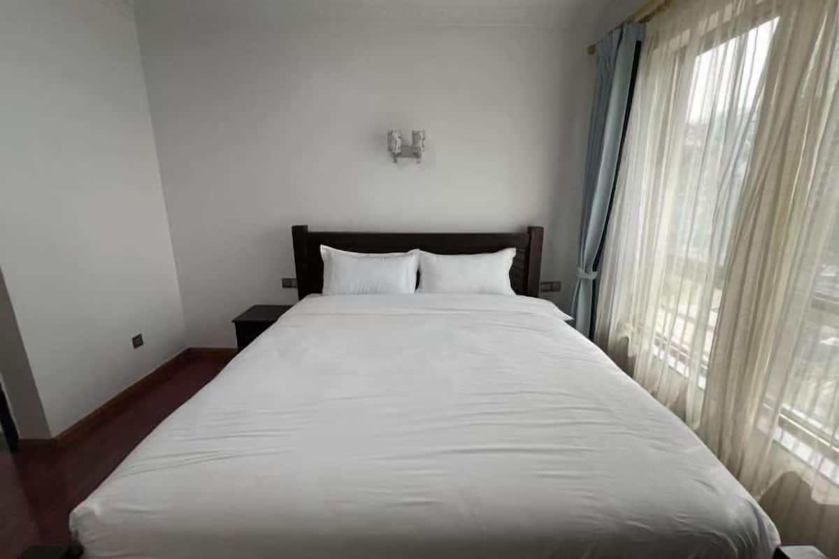 A bedroom with a large bed, a nightstand, a wall-mounted light fixture, and sheer curtains.