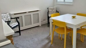 Un coin repas avec une table blanche et des chaises jaunes, un banc blanc agrémenté d’un coussin à carreaux et un lustre.
