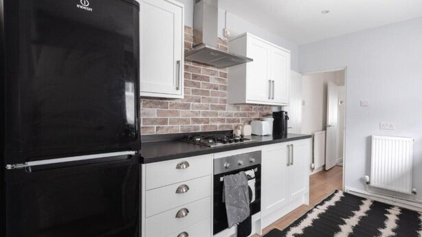 Une cuisine moderne avec un réfrigérateur noir, des armoires blanches, un mur de briques apparentes et un tapis noir et blanc.