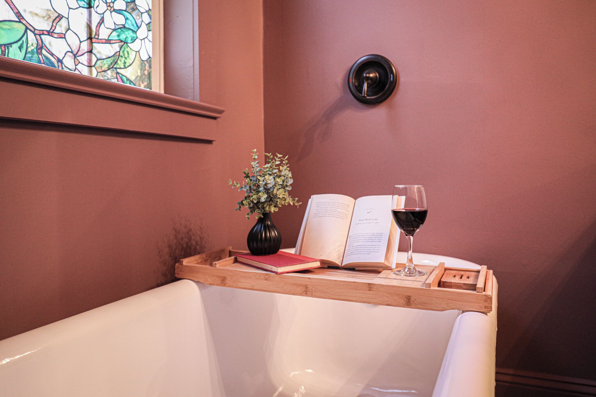 Un'area lettura con una sedia bianca, un vassoio di legno con un libro, un vaso con fiori e un bicchiere di vino rosso.