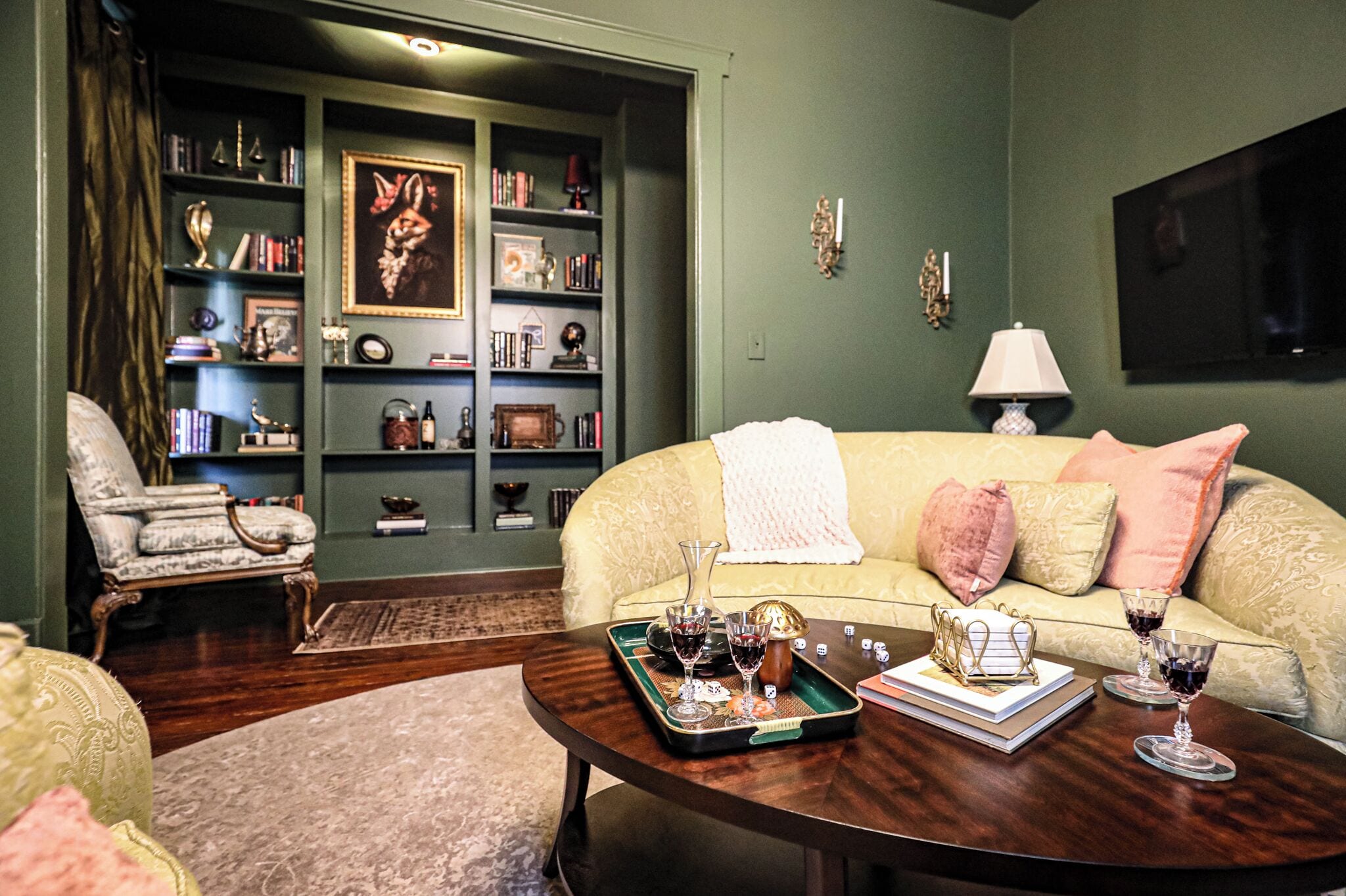 A cozy living room with a green wall, a bookshelf filled with books and decorative items, a comfortable sofa with cushions, and a coffee table with wine glasses and a tray.