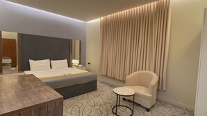 A modern hotel room with a bed, a wooden side table, a small round table, a white armchair, and a large window with vertical blinds. - Lamar Park Hotel Al Rayan (Tabuk)