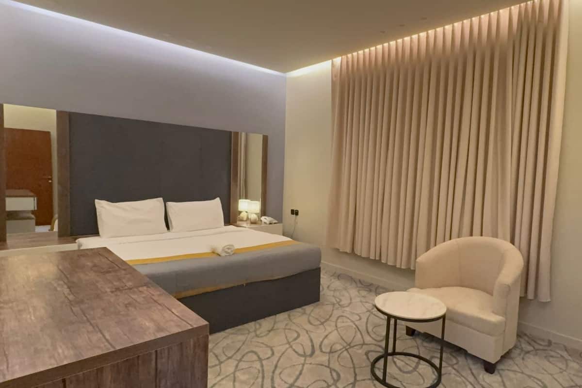 Une chambre d’hôtel moderne, équipée d’un lit, d’une table de chevet en bois, d’une petite table ronde, d’un fauteuil blanc et d’une grande fenêtre avec des stores verticaux.