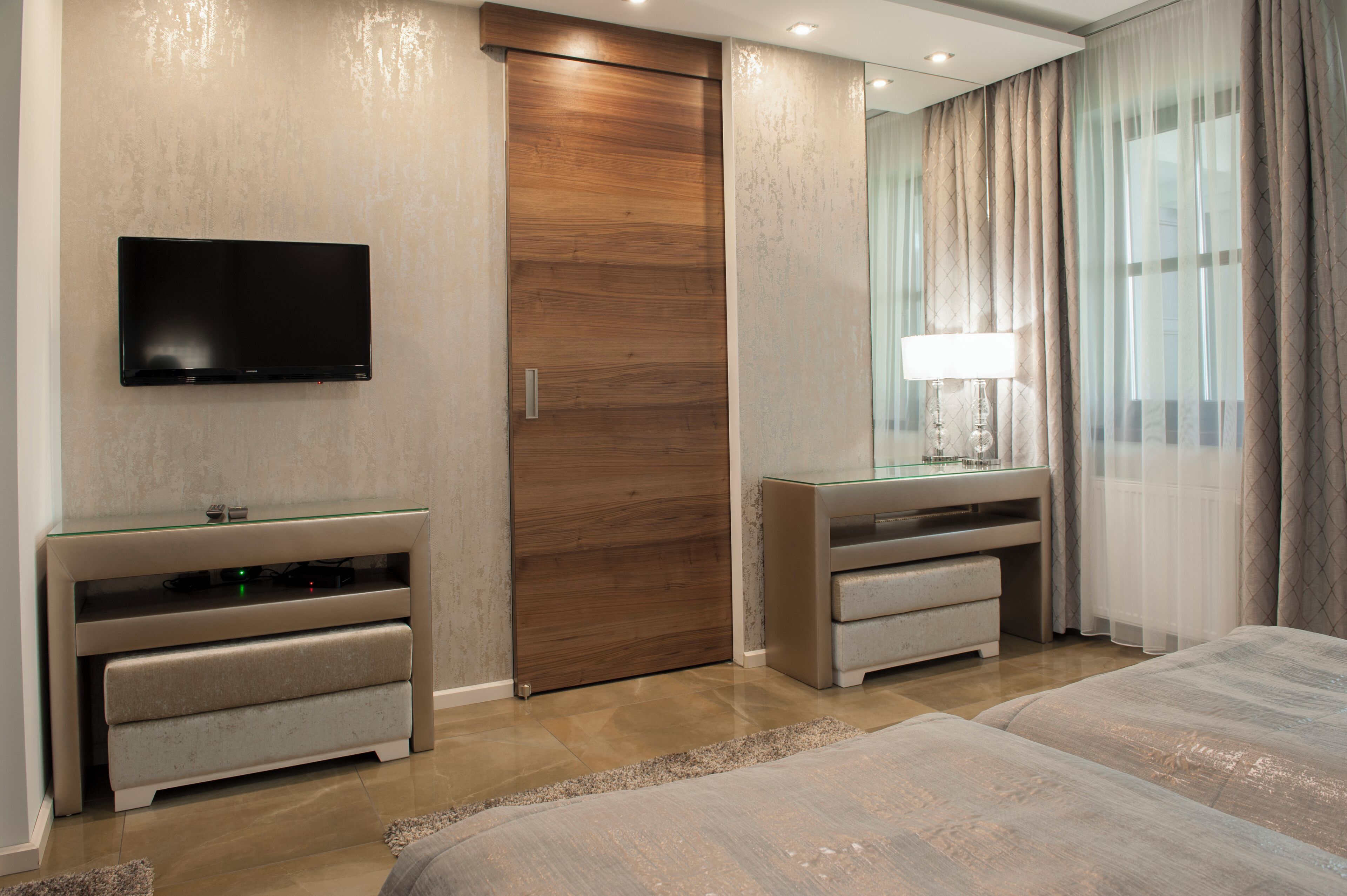 A modern hotel room with a flat-screen TV mounted on the wall, a wooden door, a glass console table with drawers, a bedside lamp, and a large window with curtains.