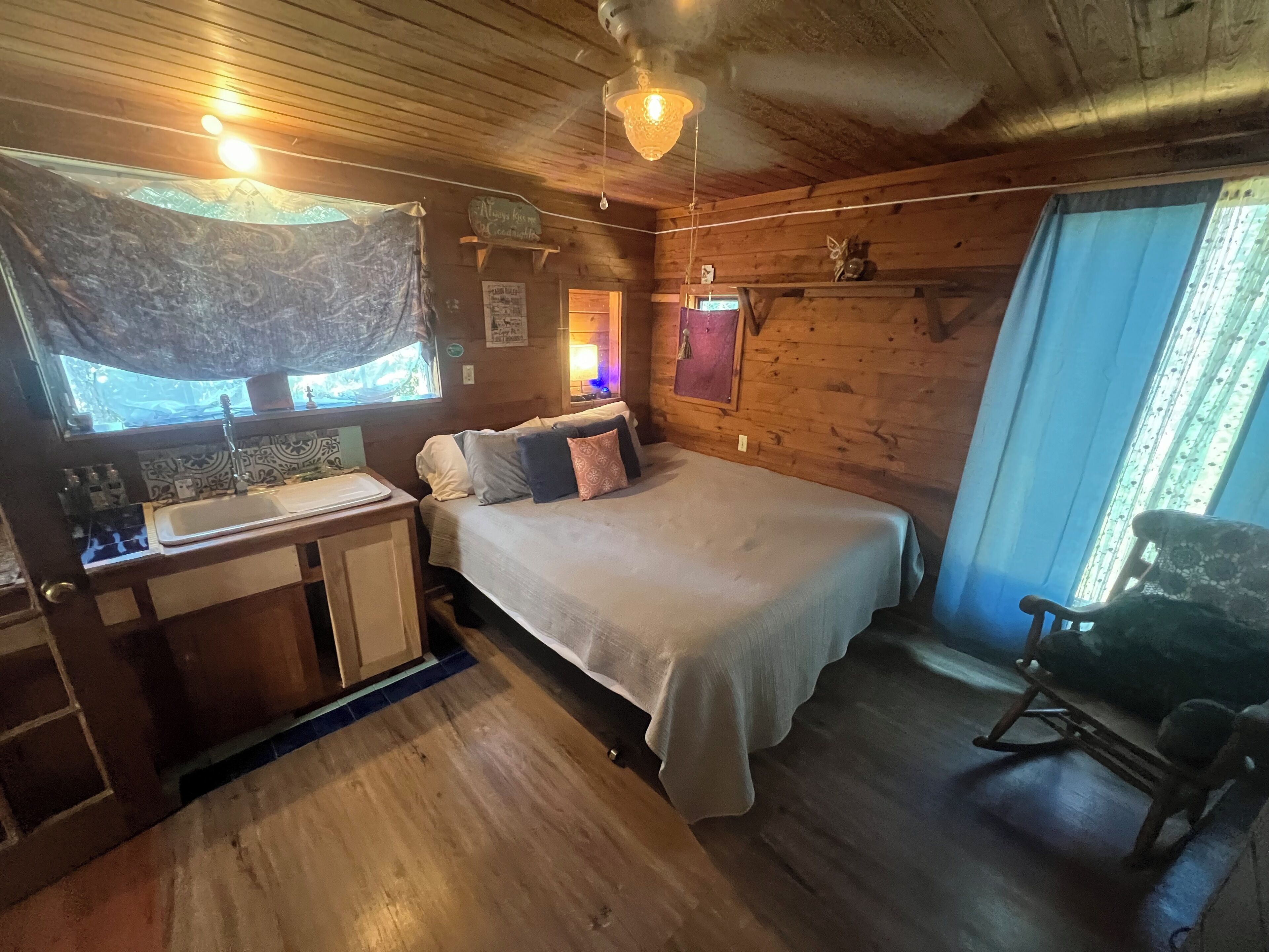 Une chambre confortable, aménagée dans le style d’un chalet, avec un lit, un petit bureau doté d’un lavabo, un fauteuil à bascule et une fenêtre avec des rideaux.