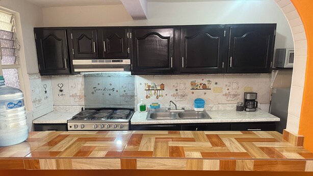 A kitchen with black cabinets, a tiled backsplash, a gas stove, a sink, and a coffee maker. There are two chairs and a large water dispenser. - Casa Vacacional Canoa 138 (La Paz)