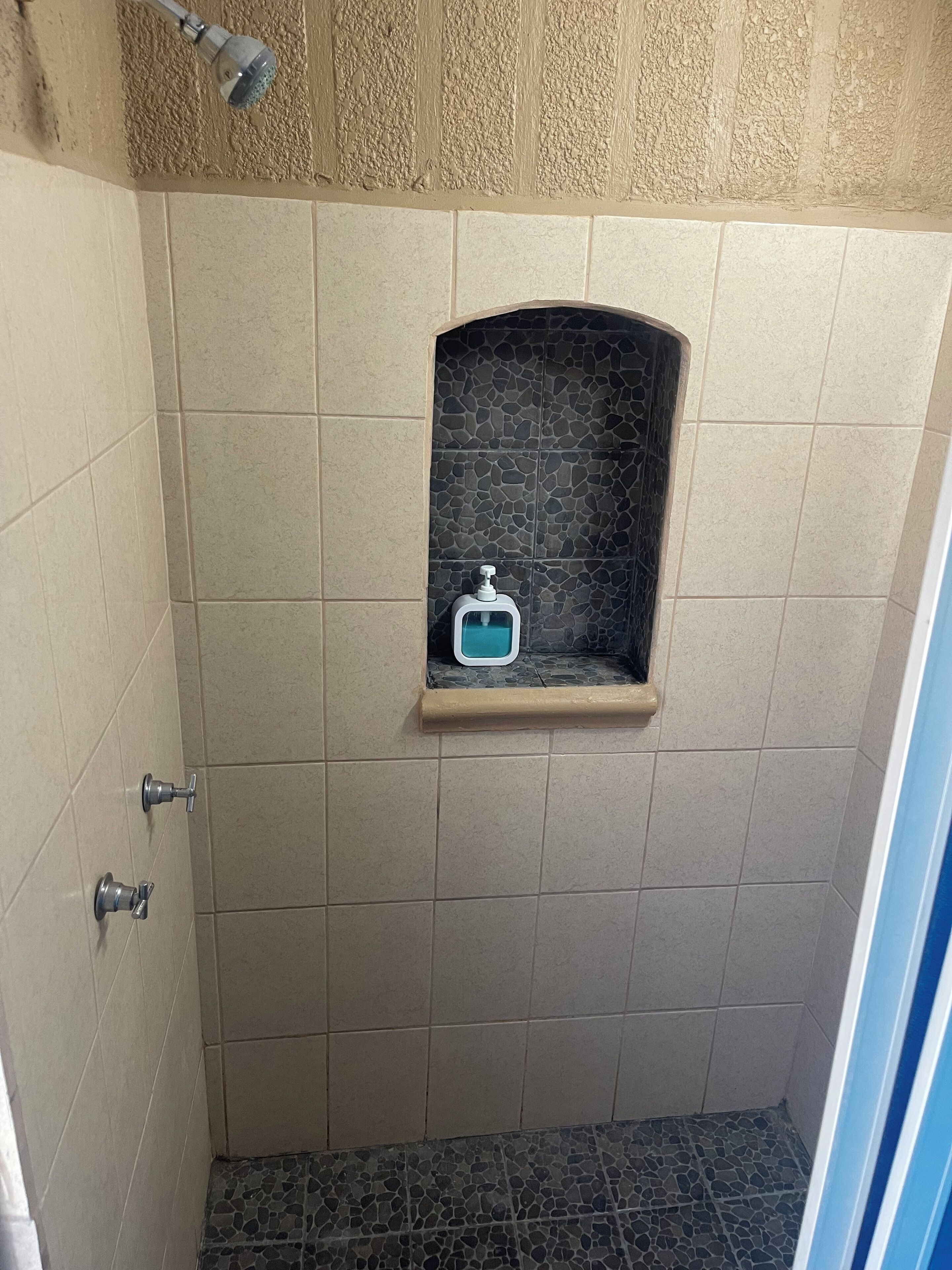 A small shower area with beige tiles, a stone-patterned niche, and a soap dispenser.