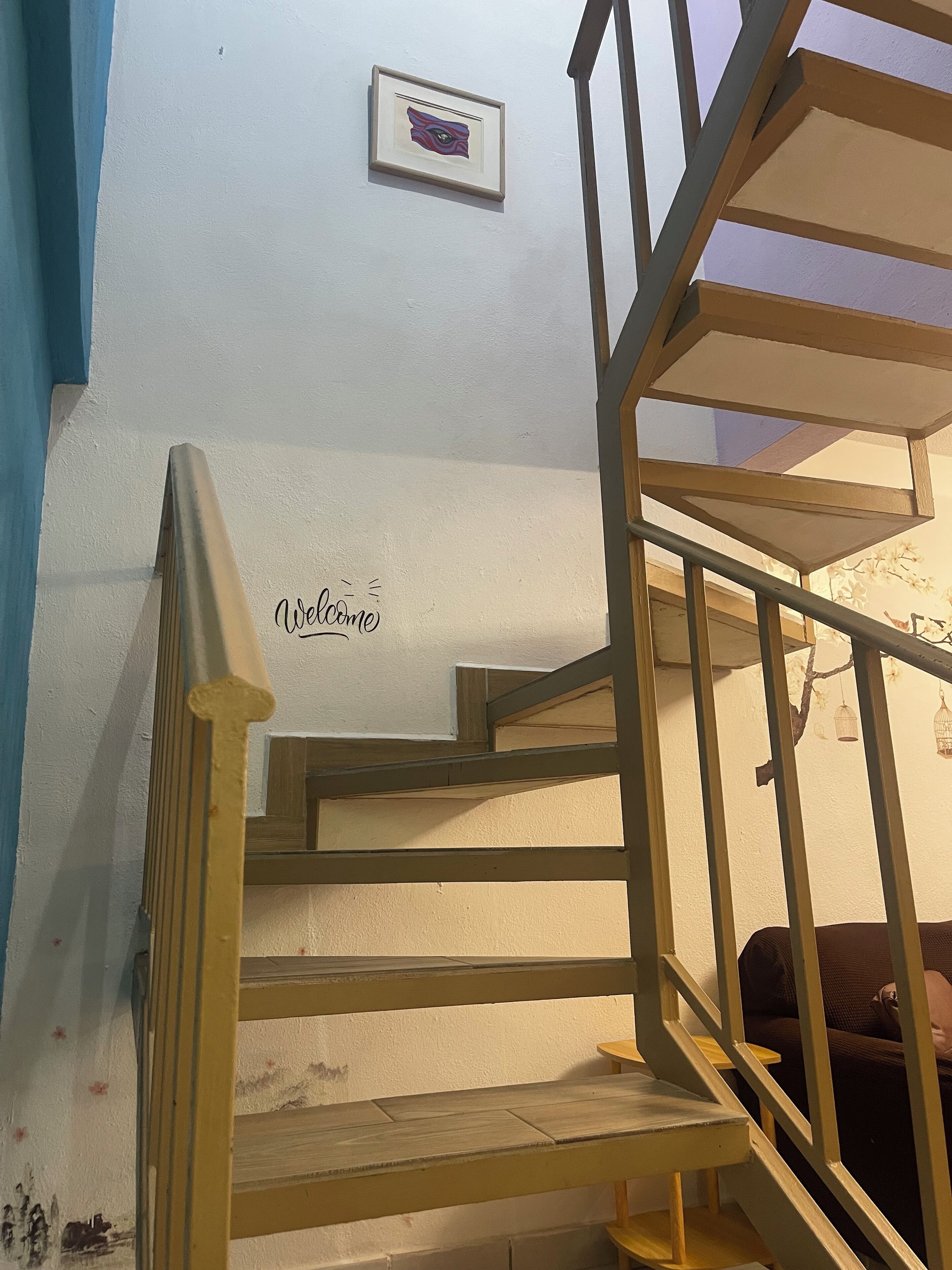 A wooden staircase with a 'Welcome' sign, a framed artwork on the wall, and a small table at the bottom.