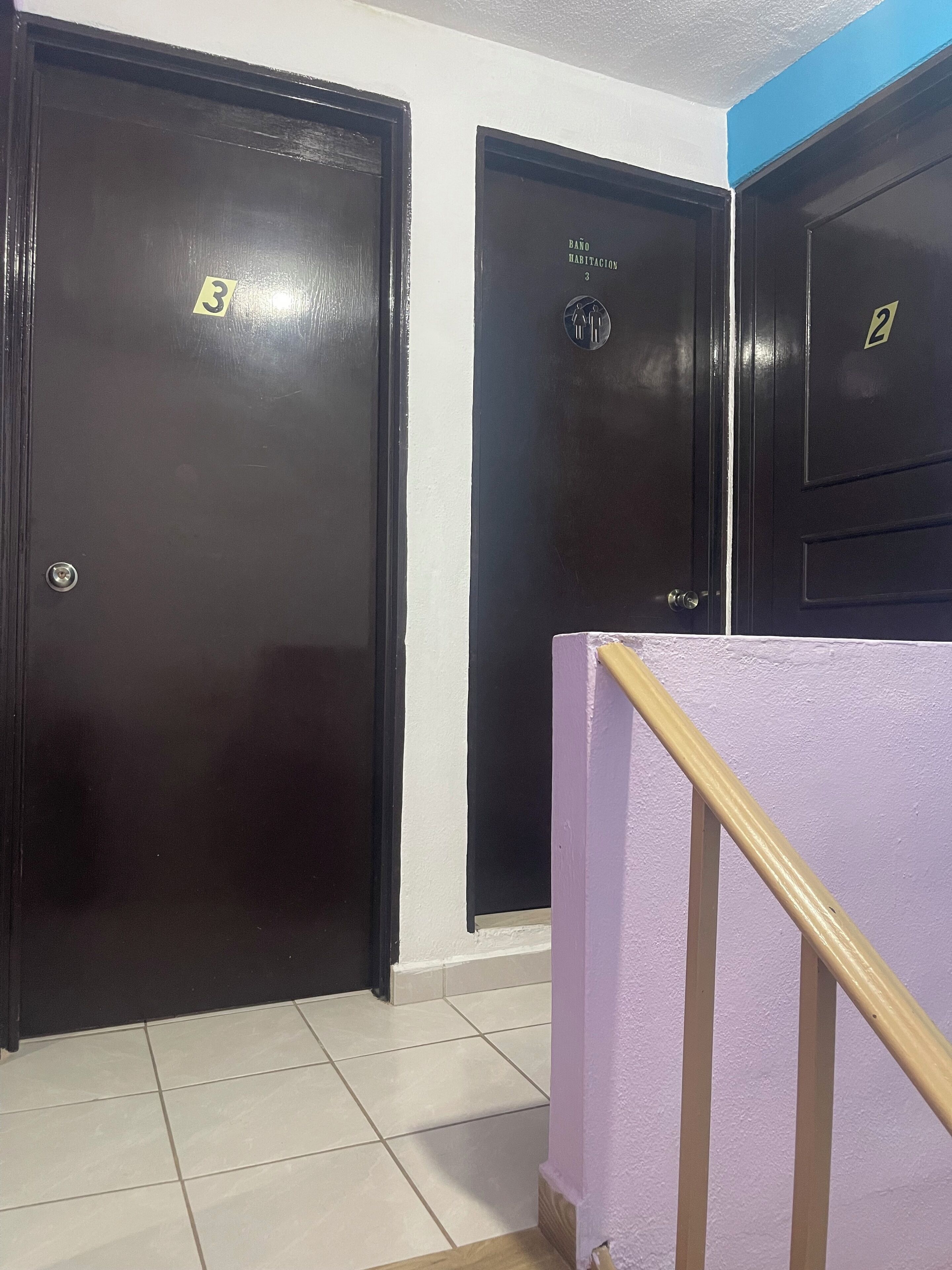 Habitación de hotel con una puerta de madera oscura, una pared blanca y una barandilla morada.