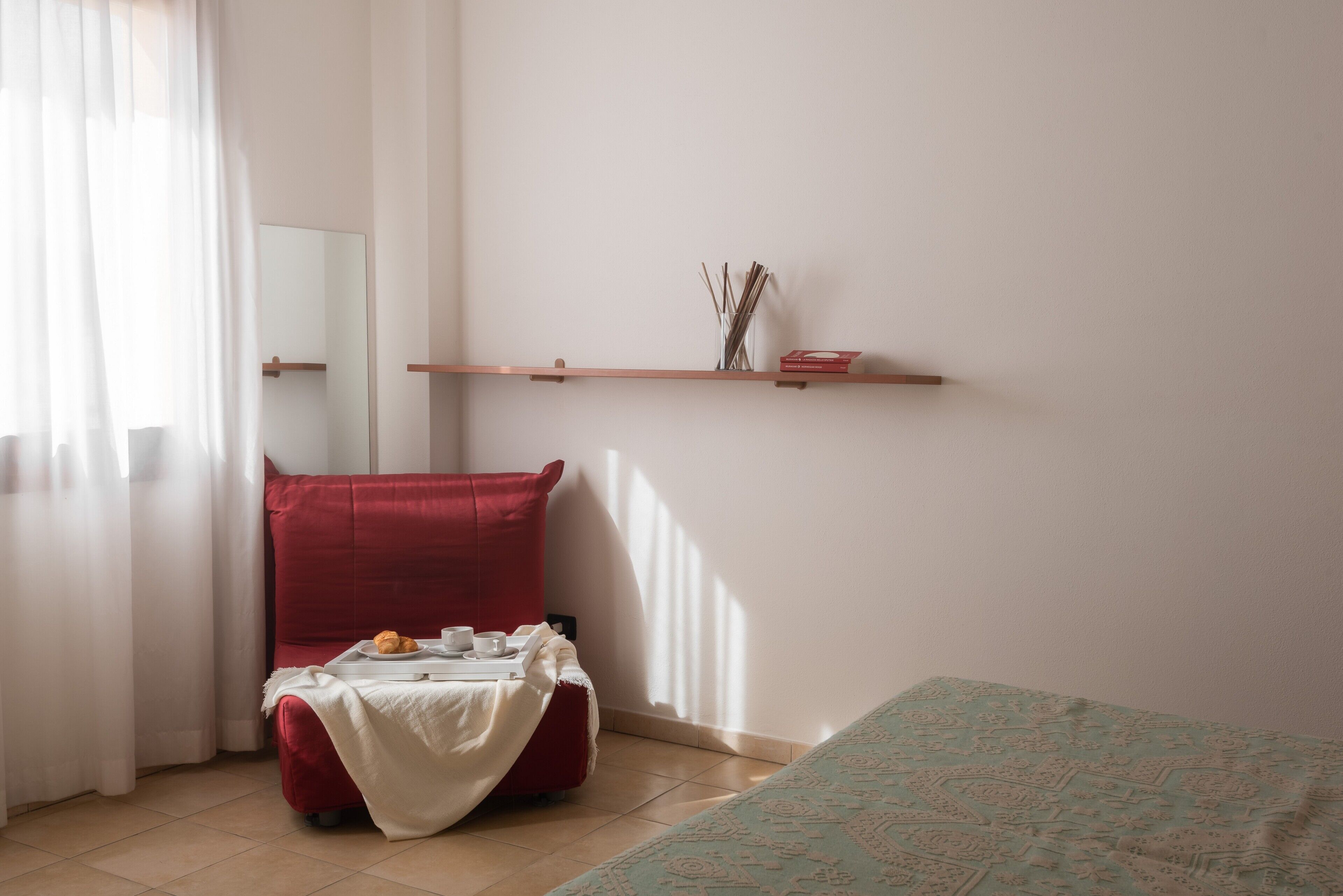 A room with a red chair, a small table with a white cloth, a shelf with decorative items, and a patterned bedspread.