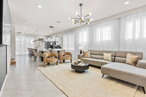 Ein modernes Wohnzimmer mit einer Eckcouch, einem runden Couchtisch und einem Essbereich mit Stühlen. Im Hintergrund ist eine Küche zu sehen.