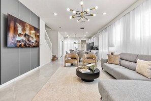 Ein modernes Wohnzimmer mit einem großen Flachbildfernseher, einem runden Couchtisch, einem grauen Sofa und einem Essbereich mit Stühlen und Tisch.