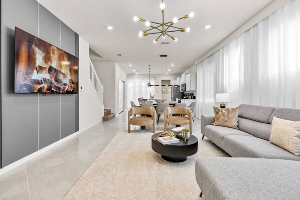 Ein modernes Wohnzimmer mit einem großen Flachbildfernseher, einem runden Couchtisch, einem grauen Sofa und einem Essbereich mit Stühlen und Tisch.