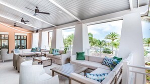 Ein überdachter Außenbereich mit Sofas und Sesseln, Deckenventilatoren und großen Fenstern mit Blick auf eine angelegte Grünfläche.
