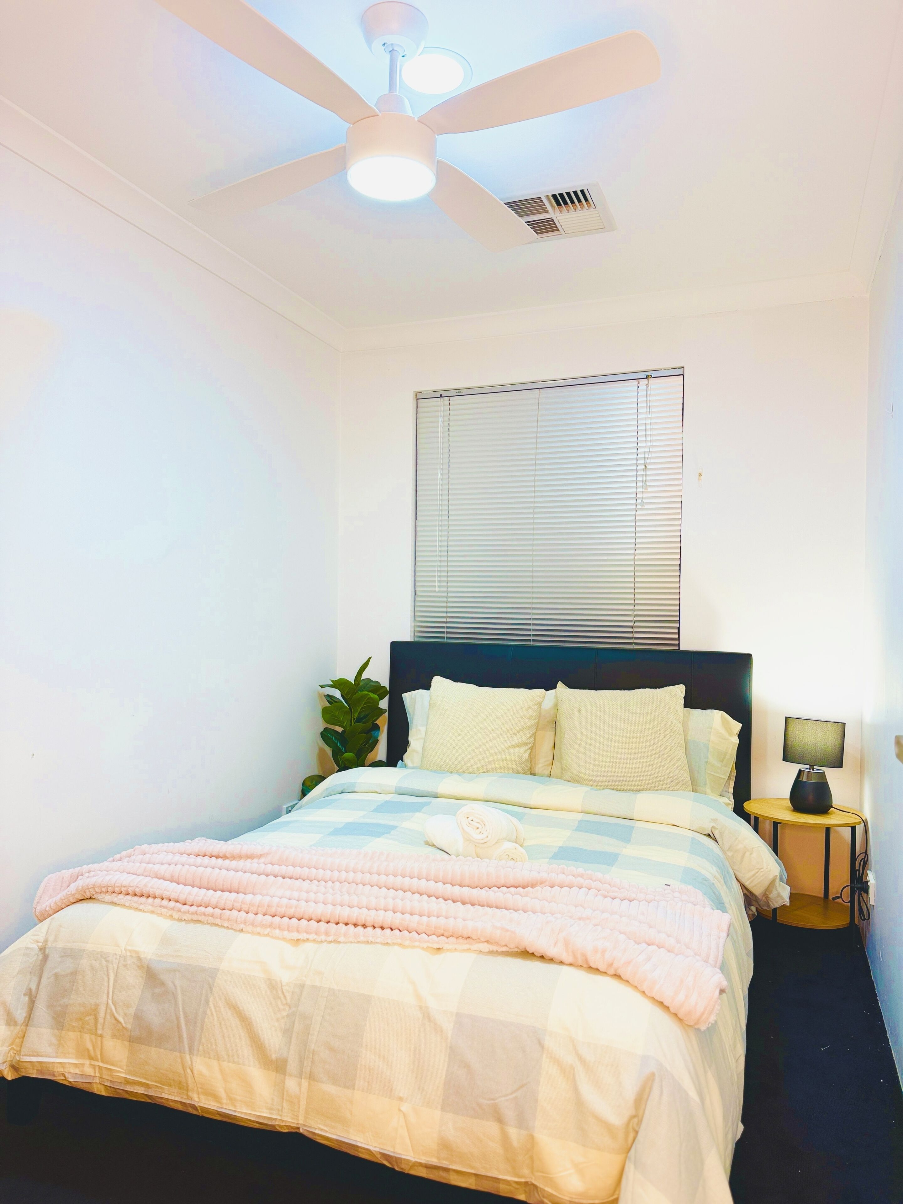 Een slaapkamer met een bed, een plafondventilator, een raam met zonwering, een nachtkastje met een lamp en een potplant.