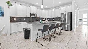Een moderne keuken met witte kastjes, een centraal eiland met barstoelen, roestvrijstalen apparatuur en een zwarte prullenbak.