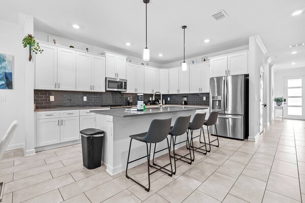 A modern kitchen with white cabinets, a central island with bar stools, stainless steel appliances, and a black trash bin. - 7BR house sleeps 17 with private pool in kissimmee (Kissimmee)