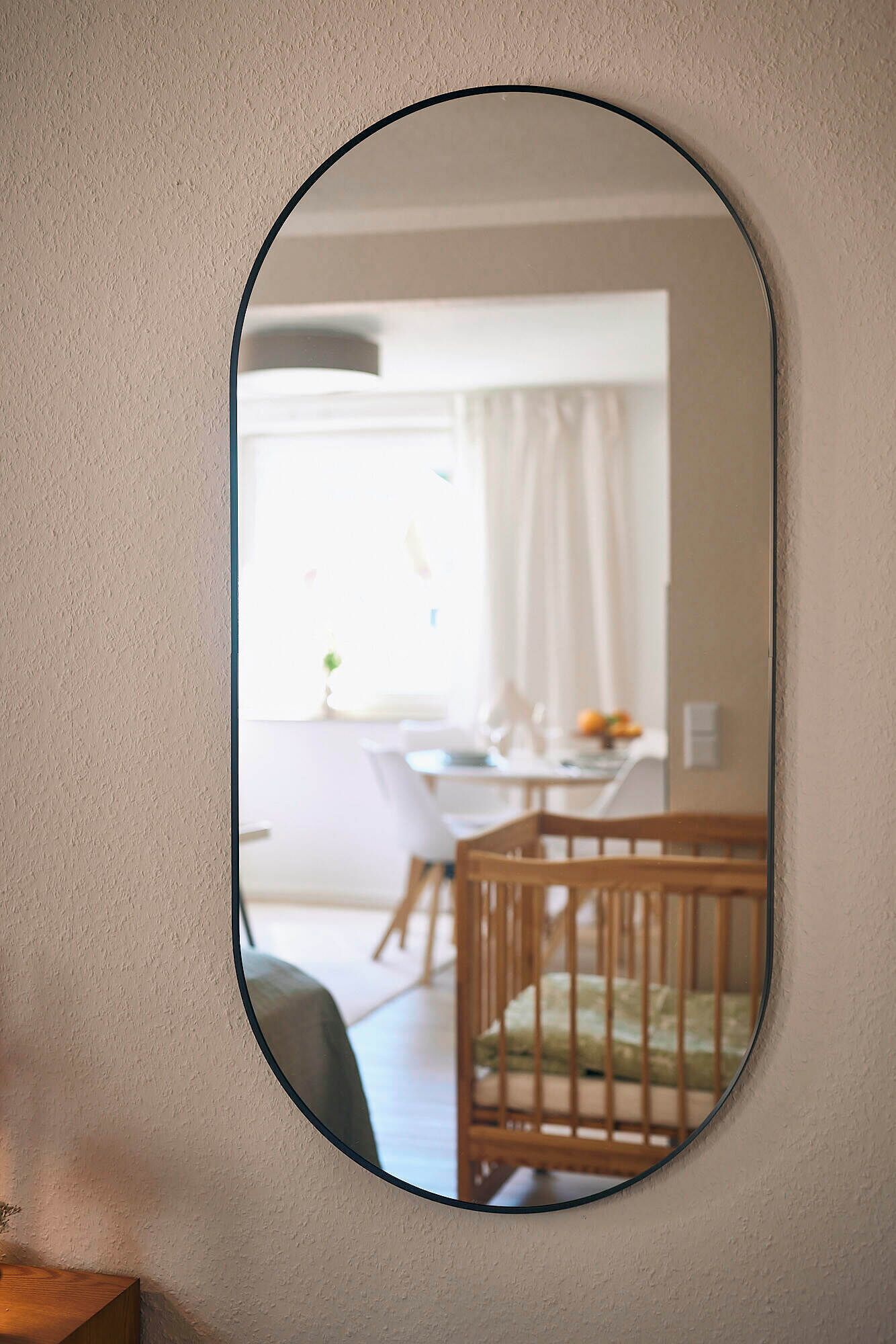Un miroir rond reflétant une pièce dans laquelle on aperçoit un berceau, une table à manger et une fenêtre avec des rideaux.