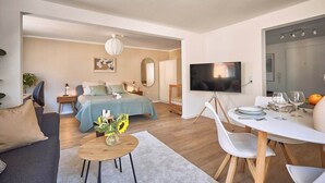 A modern, well-lit living room with a sofa, a coffee table, a dining area with a table and chairs, a TV, and a large mirror. - Cosima - Kaiserslautern (Kaiserslautern)