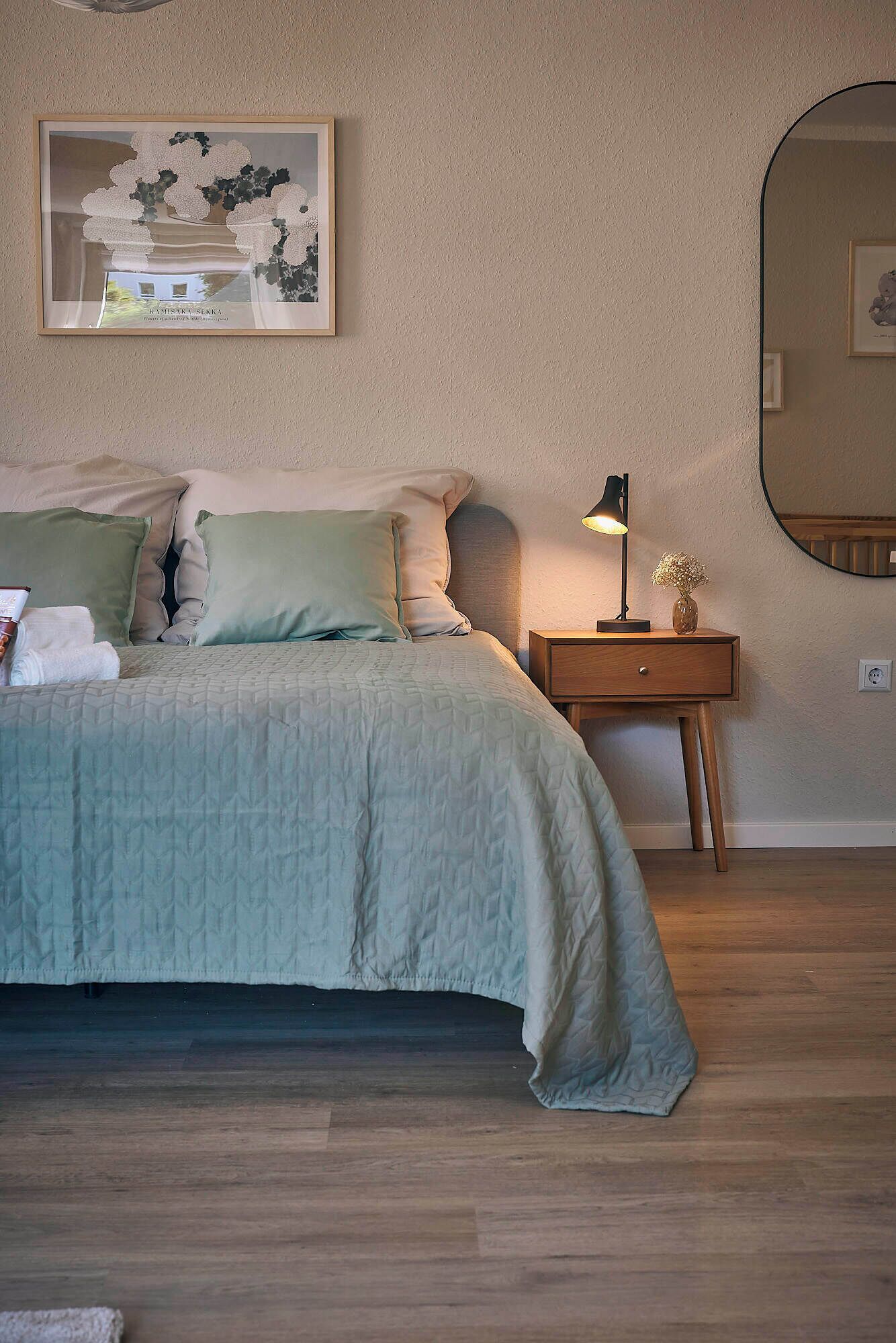 Een slaapkamer met een bed, nachtkastje, lamp, spiegel en een lijst met een afbeelding aan de muur.