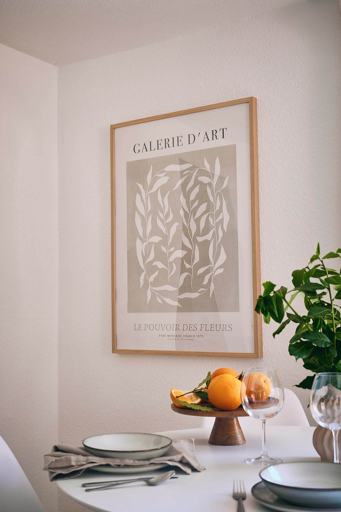 A framed artwork titled 'GALERIE D'ART' with a floral design, placed above a dining table with plates, cutlery, and a glass of water.