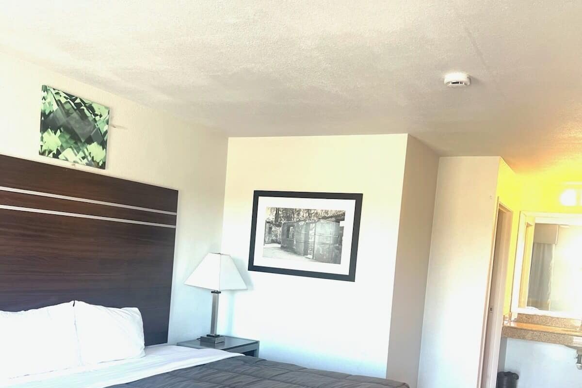 Une chambre d’hôtel avec un lit, une table de chevet, une lampe, un tableau encadré au mur et une salle de bain visible à travers une porte ouverte.