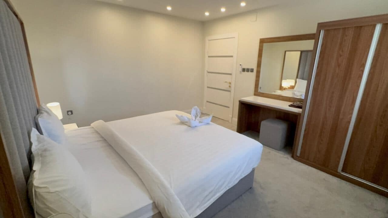 A hotel room with a neatly made bed, a wooden wardrobe, a mirror, and a small desk with a lamp.