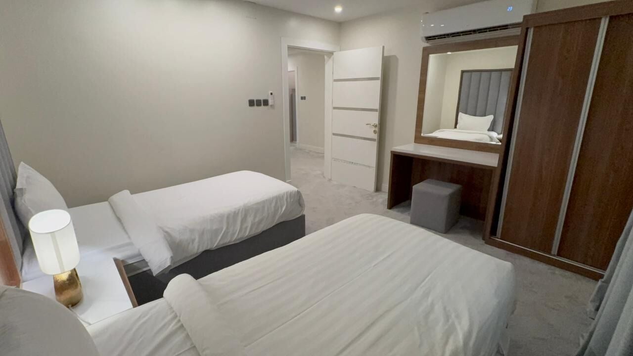 A hotel room with two beds, a desk with a lamp, a wardrobe, and a mirror.