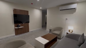 A modern hotel room with a flat-screen TV mounted on a wooden panel, a sofa, a coffee table, and a small side table with a lamp. - DIFFERENT HOTEL (Tabuk)