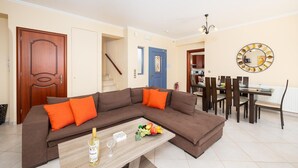 A living room with a brown sofa, orange cushions, a wooden coffee table with a bottle and glasses, and a dining area with a table and chairs.
