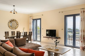 Una sala de estar con sofá, mesa de comedor, televisor sobre un mueble bajo y ventanas grandes con cortinas.